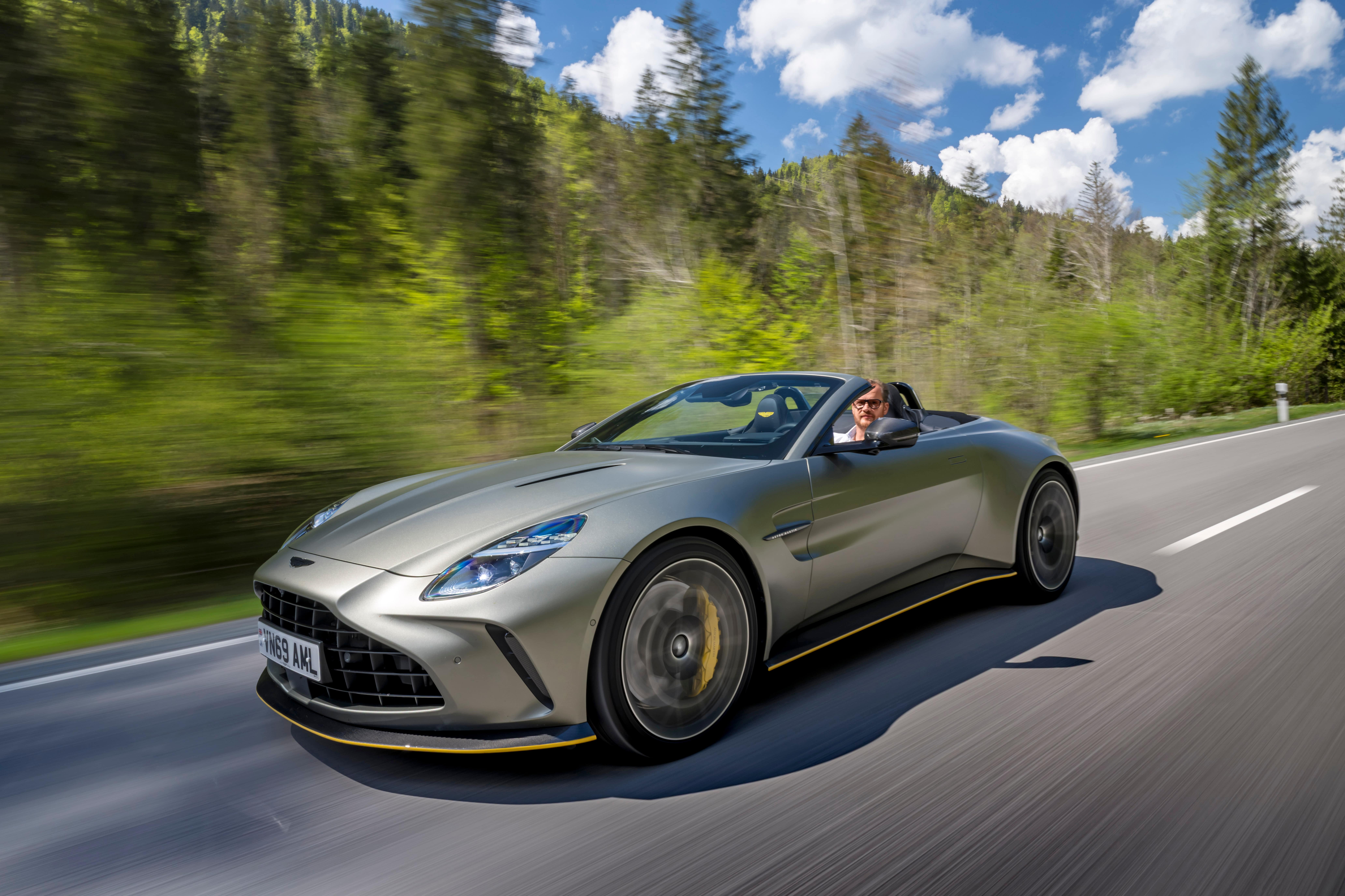 Ein silberner Aston Martin Cabrio fährt mit hoher Geschwindigkeit auf einer Landstraße durch eine grüne Waldlandschaft